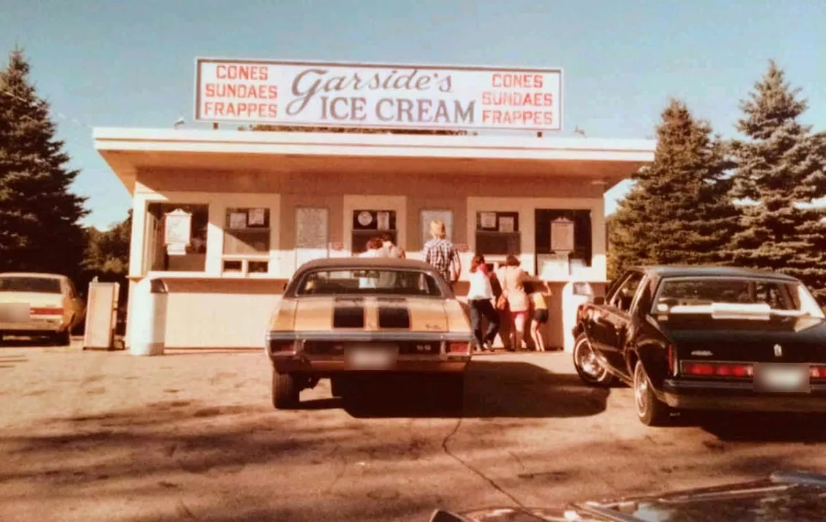 An older photo of Garside's: the stand under its red and black sign, Cones, Sundaes, Frappes, with cars of the time parked in front and customers at the windows.