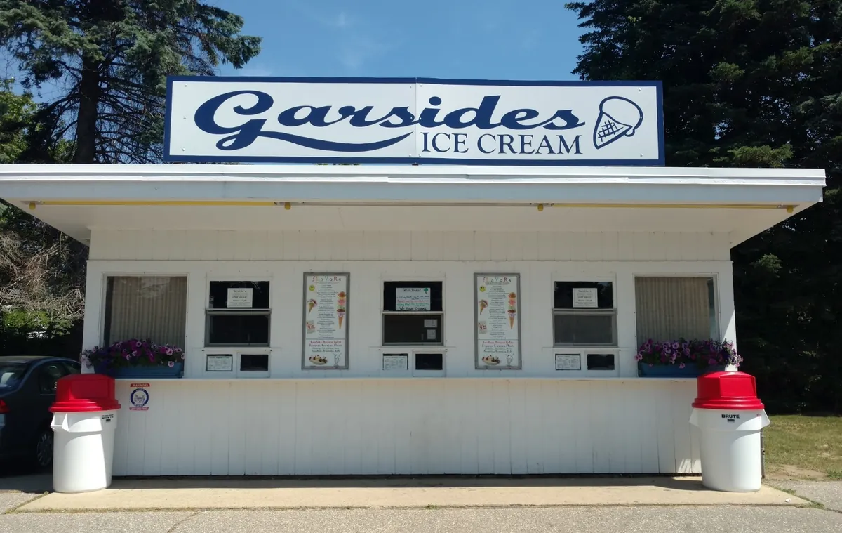 The front of Garside's: the white stand under its blue script sign, flavor menus in the windows, flower boxes and a red-topped bin at each end.