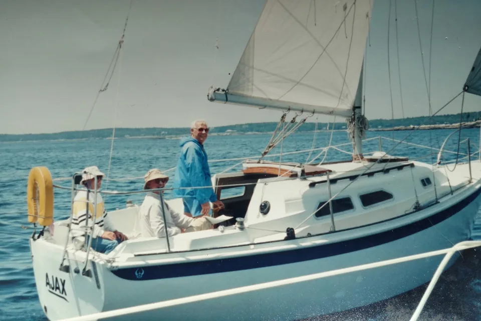 Three people aboard Ajax under sail, the man at the helm in a blue windbreaker smiling at the camera, a wooded shore behind.