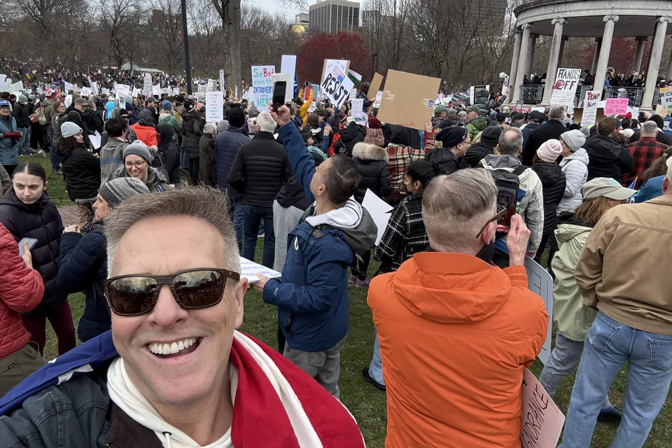 Eric grinning in sunglasses in a selfie at a crowded rally on Boston Common, signs raised behind him.