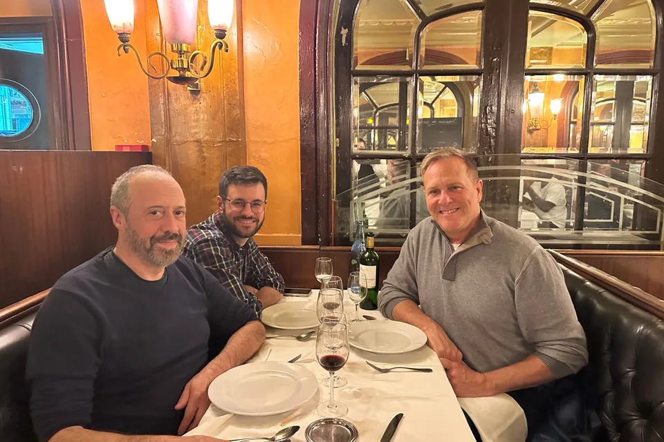 Eric and two friends at a brasserie table in Paris, wine glasses and white plates in front of them, mirrored arches behind.