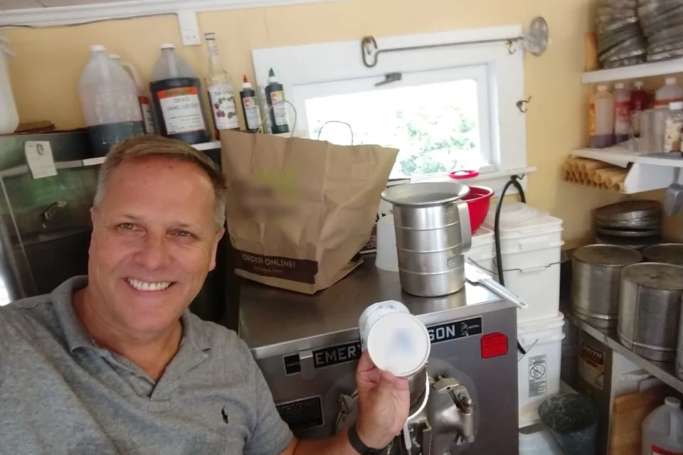 A selfie of Eric smiling inside Garside's, beside the ice cream freezer, holding up a container lid; syrups and cones on the shelves behind him.