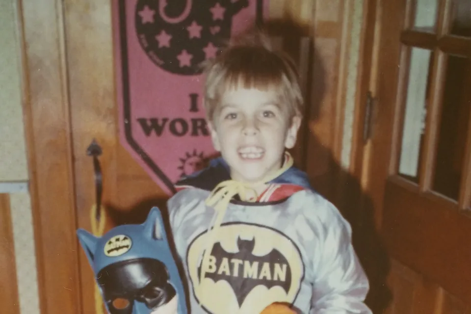 A small boy in a Batman costume holding the mask and a plastic pumpkin, grinning, on Halloween.