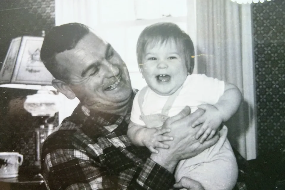 A laughing baby held up by a grinning man in a plaid shirt, in a black-and-white photo from a Maine living room.