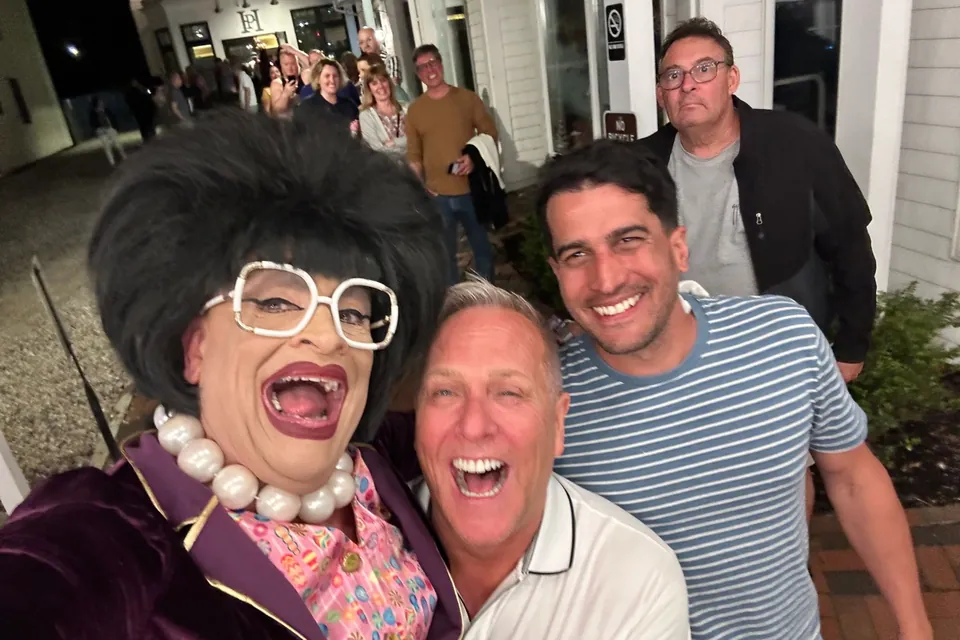 A night-time selfie in Provincetown: Eric laughing beside the comedian Miss Richfield, in her black beehive, white glasses and pearls, with friends and a crowd behind them.